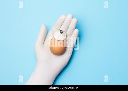 Flatlay. Hand in weißen medizinischen Handschuhen mit Ei mit Eisbeutel auf blauem Hintergrund. Pflegekonzept Stockfoto