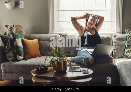 Meine eigene Firma ist meine Lieblingsfirma. Eine junge Frau, die zu Hause einen Laptop und Kopfhörer auf dem Sofa benutzt. Stockfoto