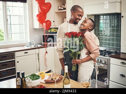 Happy Anniversary, babe. Ein liebevolles junges Paar, das mit einem Blumenstrauß in ihrer Küche zu ihrem Jahrestag posiert. Stockfoto