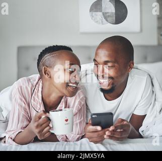 Es sind die kleinen Momente, die viel bedeuten. Ein glückliches junges Paar mit einem Smartphone und Kaffee zusammen im Schlafzimmer zu Hause. Stockfoto