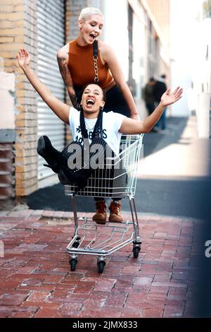 Freunde machen Trolleys Spaß. In voller Länge Aufnahme von zwei fröhlichen jungen Freundinnen, die tagsüber Spaß mit einem Einkaufswagen im Freien haben. Stockfoto
