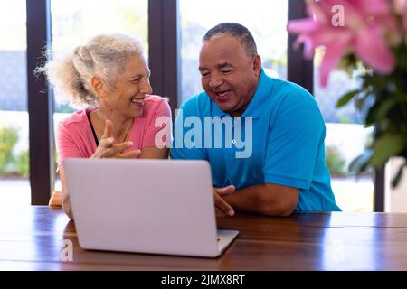 Glücklicher älterer Birazialmann und kaukasische Frau, die sich Videos über dem Laptop angucken, während sie am Tisch sitzen Stockfoto