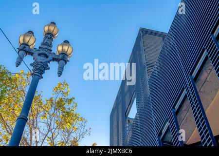 Low-Angle-Ansicht der Straßenbeleuchtung mit drei Lampen und Gebäude mit Metallwänden in San Francisco, CA. Auf der linken Seite gegenüber befindet sich ein Laternenpfosten und ein Baum Stockfoto