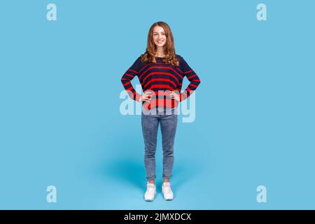 Porträt einer positiven glücklichen Frau in einem gestreiften Pullover im lässigen Stil und Jeans, die mit den Händen auf den Hüften stehen und die Kamera betrachten. Innenaufnahme des Studios isoliert auf blauem Hintergrund. Stockfoto