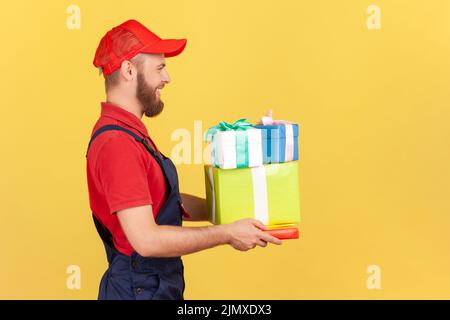 Seitenansicht Porträt eines positiven glücklichen Kuriermanns in Uniform und Mütze mit vielen Geschenk-Boxen, die Ordnung für die Feier des Urlaubs bringen. Innenaufnahme des Studios isoliert auf gelbem Hintergrund Stockfoto