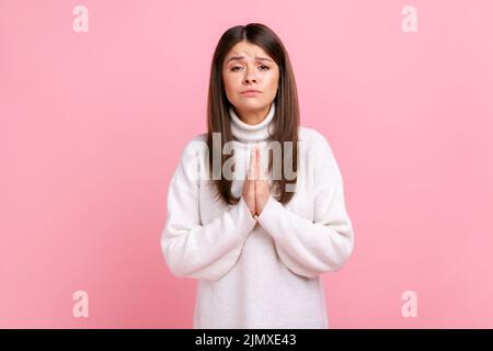 Das Weibchen hält die Hände im Gebet und sieht mit flehenden Bitten, bettelender Hilfe, Vergebung aus, trägt einen weißen Pullover im lässigen Stil. Innenaufnahme des Studios isoliert auf rosa Hintergrund. Stockfoto