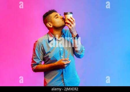 Porträt eines hübschen jungen erwachsenen Mann im Hemd küssen Einweg-Tasse Kaffee zum Mitnehmen, genießen Getränk, braucht Energie. Innenaufnahmen im Studio, isoliert auf farbigem Neonlicht-Hintergrund. Stockfoto