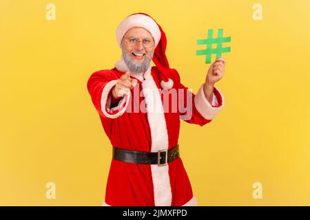 Positiver älterer Mann mit grauem Bart, trägt ein weihnachtsmann-Kostüm und steht mit einem grünen Hashtag in den Händen, der mit einem toothy Lächeln auf die Kamera zeigt. Innenaufnahme des Studios isoliert auf gelbem Hintergrund. Stockfoto