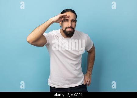 Porträt eines neugierigen Mannes mit Bart, der ein T-Shirt trägt, die Handfläche über dem Kopf hält und aufmerksam weit weg blickt und mit Erwartung auf weite Strecken blickt. Innenaufnahme des Studios isoliert auf blauem Hintergrund. Stockfoto