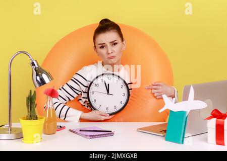 Traurige verärgert Frau in Gummiring am Hals sitzen am Arbeitsplatz mit Laptop, zeigt Wanduhr, hat keine Zeit, in den Sommerurlaub zu gehen. Studio-Innenaufnahme isoliert auf gelbem Hintergrund. Stockfoto