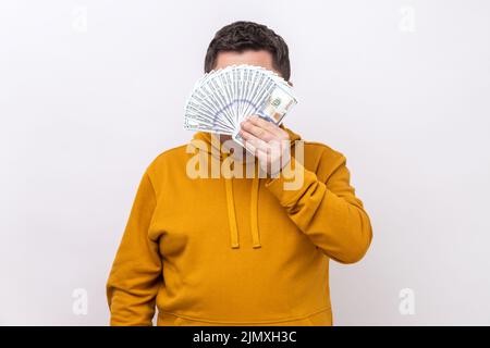 Unbekannter Mann versteckt sich hinter einem Fan von hundert Dollar-Scheinen, großer Gewinner, Wetten, trägt einen Kapuzenpullover im urbanen Stil. Innenaufnahme des Studios isoliert auf weißem Hintergrund. Stockfoto