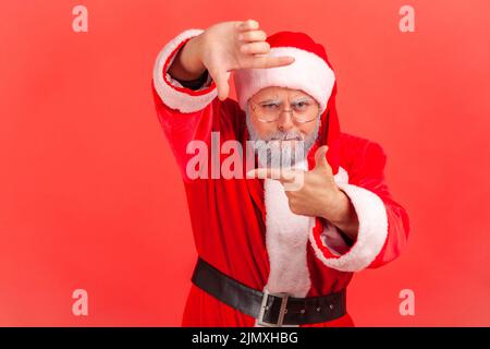 Älterer Mann im weihnachtsmann-Kostüm gestikulierend Bilderrahmen mit Händen, durch die Finger schauen und sich auf interessante Momente konzentrieren, fotografieren. Innenaufnahme des Studios isoliert auf rotem Hintergrund. Stockfoto