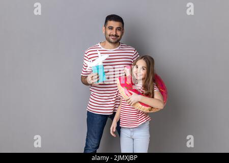 Porträt von fröhlichen glücklichen Vater und Tochter in gestreiften T-Shirts werden Berufung haben, halten Gummiring, Pass und Papierflugzeug. Innenaufnahme des Studios isoliert auf grauem Hintergrund. Stockfoto