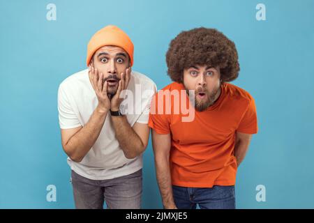 Das Porträt zweier erstaunter junger, erwachsener Hipster-Männer, die mit großen Augen und offenem Mund die Kamera betrachten, ist schockiert von Nachrichten und sieht etwas Erstaunliches. Innenaufnahme des Studios isoliert auf blauem Hintergrund. Stockfoto