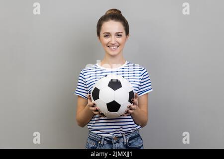 Porträt einer Frau mit gestreiftem T-Shirt, die mit einem angenehmen Lächeln auf die Kamera blickt, den Fußball hält und Fußballspiele anschaut. Innenaufnahme des Studios isoliert auf grauem Hintergrund. Stockfoto