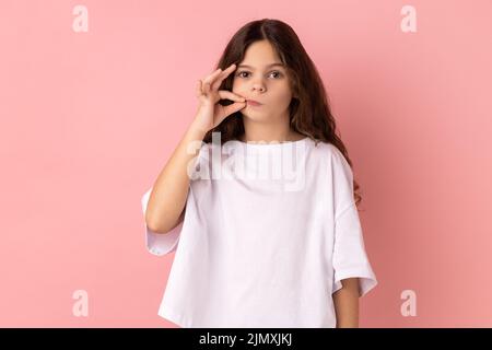 Portrait von charmanten kleinen Mädchen trägt weiße T-Shirt mit Mystery-Look macht Reißverschluss Geste zu schließen Mund, geheim zu halten, Zippen Lippen. Innenaufnahme des Studios isoliert auf rosa Hintergrund. Stockfoto