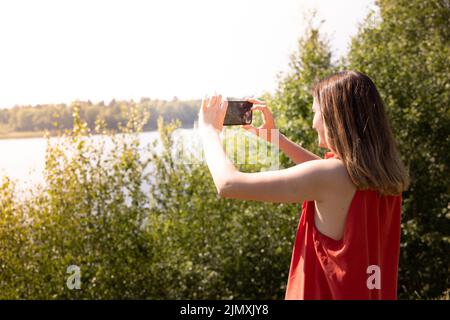 Die hübsche junge Kaukasierin im roten Rock macht Fotos mit dem Handy im grünen Wald am See draußen Stockfoto