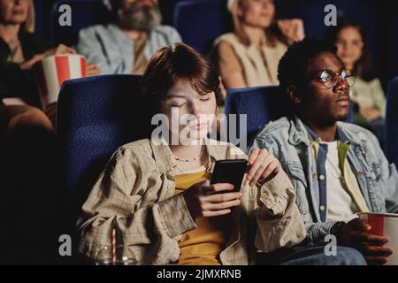 Junge Frau der Generation Z Kaukasische langweilt sich im Kino mit dem Film-Scrolling-News-Feed in einem sozialen Netzwerk auf dem Smartphone Stockfoto