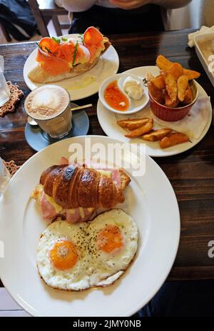 Nahaufnahme geräucherter Lachs auf Toast, serviert mit Kartoffelscheiben, Schinken-Käse-Croissant und heißem Latte-Kaffee Stockfoto