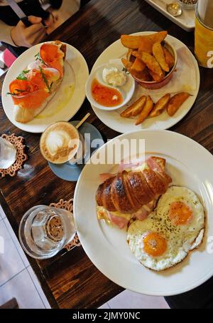 Nahaufnahme geräucherter Lachs auf Toast, serviert mit Kartoffelscheiben, Schinken-Käse-Croissant und heißem Latte-Kaffee Stockfoto