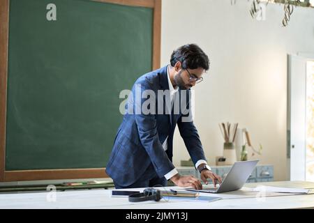 Junger indischer Geschäftsmann, Lehrer, der auf einem Laptop im Büro im Klassenzimmer steht. Stockfoto