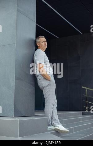 Porträt eines müden grauen Geschäftsmannes, der sich auf die graue Konstruktion eines modernen Bürogebäudes in der Nähe des Eingangs stützt. Urbaner Lifestyle, stylisch Stockfoto