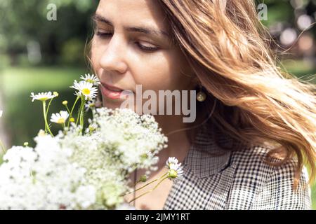 Portrait Nahaufnahme einer jungen charmanten Frau, die nach weißen Wildblumen riecht und genießt. Spaziergang im Park an sonnigen Tag, im Freien Stockfoto