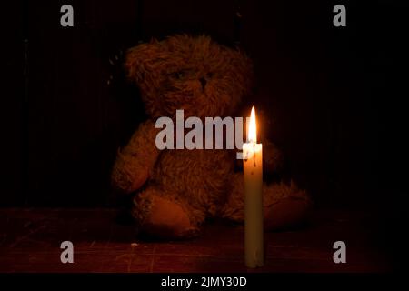 Ein brauner Teddybär im Dunkeln sitzt neben einer Kerze, ein Kinderzimmer im Dunkeln, Angst und Schrecken Stockfoto