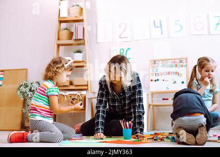 Kinder und Pädagogen falten lego und zeichnen mit Buntstiften und Markern sitzen auf dem Boden im Spielzimmer von unten sehen. Interessante Lektion für Stockfoto