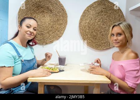 Junge Frauen blicken auf die Kamera und lächeln beim gemeinsamen Frühstück in einem Restaurant. Freundschafts- und Essenskonzept. Stockfoto