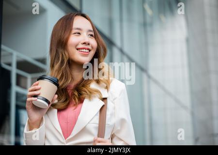 Glückliche Geschäftsfrau lächelnd halten Papiertasse mit heißem Getränk im Freien auf der Straße, asiatische Geschäftsfrau hält Kaffeetasse zum Mitnehmen gehen sie zur Arbeit Stockfoto