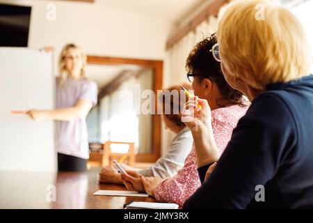 Rückansicht von drei älteren Frauen, die am Schreibtisch sitzen, Notizen machen und dem Dozenten zuhören. Sprecher erklärt die Strategie. Stockfoto