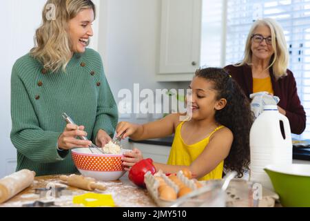 Multirassische ältere Frau, die glückliche Tochter und Enkelin beim Mischen von Essen in einer Schüssel in der Küche ansieht Stockfoto