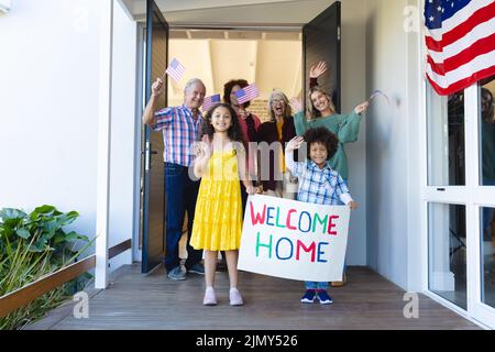 Multirassische, fröhliche, mehrerdige Familie mit Willkommenstext und amerikanischen Flaggen am Eingang Stockfoto