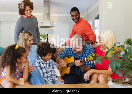 Multirassische glückliche Familie mit mehreren Generationen, die einen älteren Mann beim Gitarrenspielen im Wohnzimmer ansieht Stockfoto