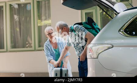 Ältere Menschen beladen Fahrzeug mit Gepäck und Trolleys während der Hochzeit Jubiläum citybreak. Seniorehepaar, das in den Urlaub in den Ruhestand geht, während es Reisegepäck in den Kofferraum des Autos legt Stockfoto