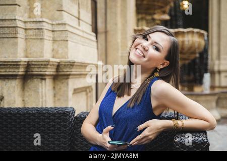 Elegante asiatische junge Frau mit einem Smartphone in den Händen lächelnd, während sie in einem Café im Freien sitzt Stockfoto