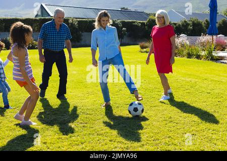 Glückliche multirassische Familie, die an sonnigen Tagen Fußball auf Gras im Hof spielt Stockfoto