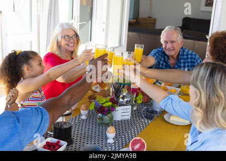 Blick aus der Perspektive auf fröhliche, multirassische Familien mit mehreren Generationen, die Saftgläser am Esstisch toasten Stockfoto