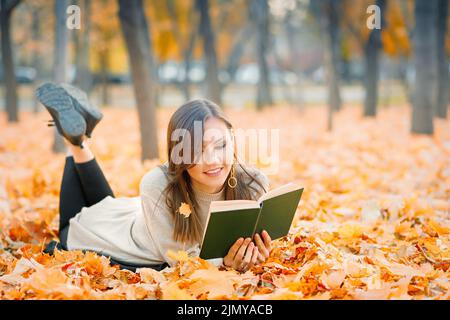 Frau mit gemischter Rasse, die auf Herbstlaub liegt und im Herbstpark ein Buch liest Stockfoto