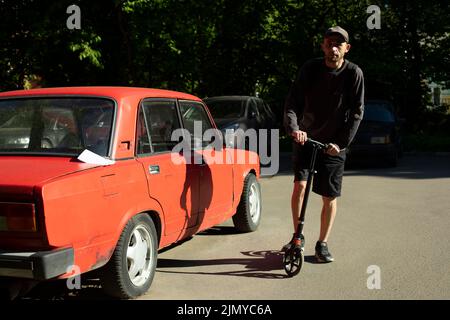 Mann auf dem Roller auf dem Parkplatz. Mann in der Nähe des roten Autos. Parkdetails. Mann in der Stadt im Sommer. Stockfoto