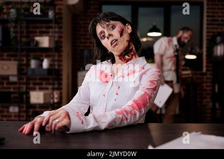 Porträt eines bösen Zombies mit Wunden im Büro, am Schreibtisch sitzend, um auf dem Laptop zu arbeiten, erschreckend und gruselig. Grausam besessen Gehirn essen Monster mit blutigem Mund offen, zu Fuß tot Teufel. Stockfoto