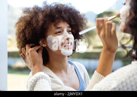 Lächelnde Biracial Freundinnen, die Schönheitscreme auf die anderen Gesichter auf dem Balkon auftragen Stockfoto