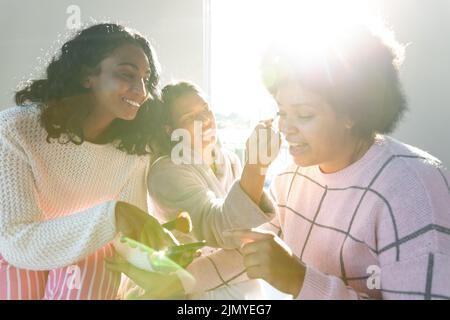 Biracial glückliche Freunde, die Make-up und zeigen Handy an junge Frau gegen Fenster Stockfoto