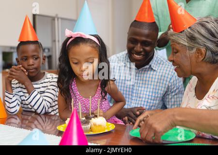 Multirassische aufgeregt Mädchen trägt Hut weht Kerzen während der Feier Geburtstag mit Familie zu Hause Stockfoto