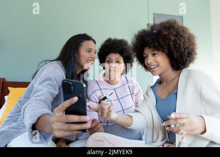 Biracial lächelnde junge Frau zeigt Smartphone aufgeregt weiblichen Freunden mit afro Haar auf dem Bett Stockfoto