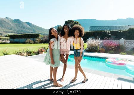 Porträt von Biracial Freundinnen lachen, posieren und hängen am Pool gegen klaren Himmel Stockfoto