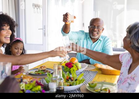 Mehrrassiger älterer Mann, der beim Mittagessen mit einer Familie aus mehreren Generationen am Esstisch Toast anhob Stockfoto