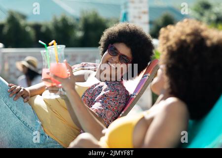Glückliche, jahrtausendealte Biracial-Freundinnen mit afro-hair-Toasting-Getränken, während sie sich auf Liegestühlen entspannen Stockfoto
