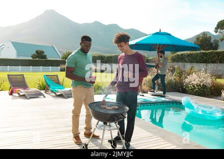 Männliche multirassische Freunde, die im Sommer Bier trinken und Fleisch auf dem Grill am Pool kochen Stockfoto